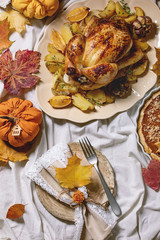 Thanksgiving or Halloween dinner with baked chicken with potatoes and lemons on big ceramic dish, pumpkin pies, plates, yellow autumn leaves, pumpkins as decorations over white table cloth. Flat lay