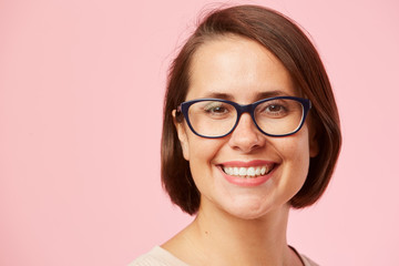 Portrait of young happy woman with short brown hair and in eyeglasses smiling at camera over pink background