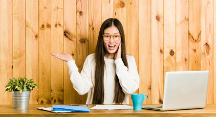 Young chinese woman studying on her desk holds copy space on a palm, keep hand over cheek. Amazed and delighted.