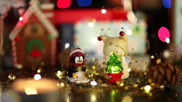 Christmas toys - cute penguin and a little snowman holding small luminous christmas tree are in the foreground. Golden sequins fall down like rain. Defocused colorful lights behind the scene.