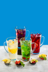 Group of lemonade jars with different ingredients and sorts of beverage at concrete textured table decorated with fruits and berries isolated at blue background.