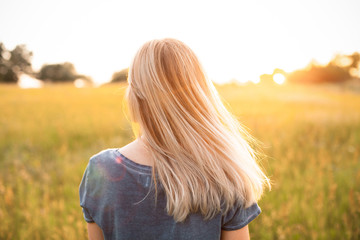 Young woman with blonde hair standing on the field and looking on the sunset