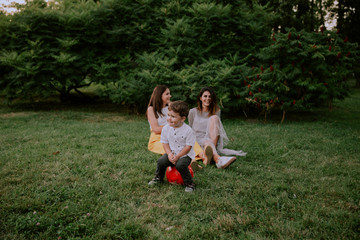 Fototapeta premium Very cute two years boy make funny faces in front of the camera while on the background two ladies smiling large and enjoying the time spending in the park