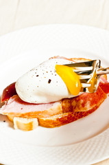 Top, front view, close up of a fork piercing a soft boiled egg showing the warm running yolk on top of a slice of fried ham on white toast, on a white, round plate