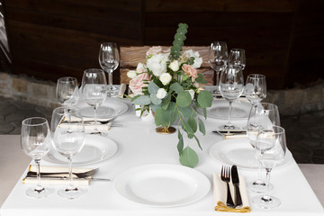 Wedding table setting decorated with fresh flowers in a brass vase. Wedding floristry. Banquet table for guests outdoors with a view of green nature. Bouquet with roses, eustoma and eucalyptus leaves