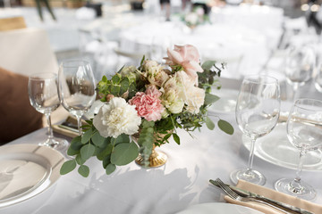Wedding table setting decorated with fresh flowers in a brass vase. Wedding floristry. Banquet table for guests outdoors with a view of green nature. Bouquet with roses, eustoma and eucalyptus leaves