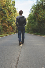 A young man with a backpack behind him is walking along an empty autumn road. Travel, hipster. Walk in the fall, autumn trees.