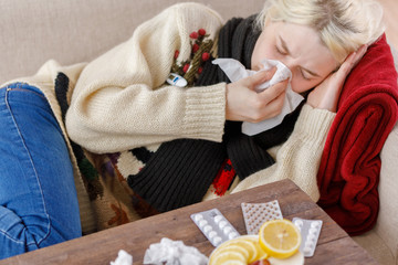 Girl in a sweater sneezes while sitting on a sofa. Colds and flu. The patient caught a cold,...