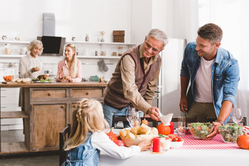 selective focus of smiling father and grandparent looking at kid and setting table in Thanksgiving day