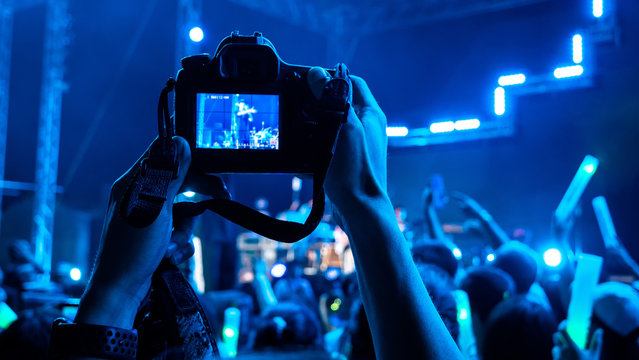 Photographer taking picture with camera at a festival concert with crowd people raised hands and attending a concert. Blue black light by stage lights. Summer music festival concept.