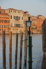 Wooden columns of a mooring and a lantern, water of the Grand Canal and buildings of Venice. Light from the setting sun
