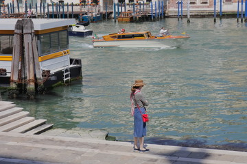 Fototapeta na wymiar A girl in a hat stands with her back (half-turned) and looks at the Grand Canal in Venice. A taxi boat sails past.
