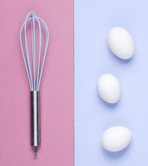Kitchen whisk, chicken eggs on pastel colored background. Top view