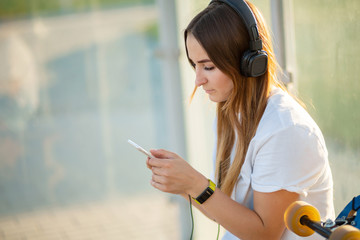 Stylish young girl sitting on bus stop with her longboard, listening music and wait for bus