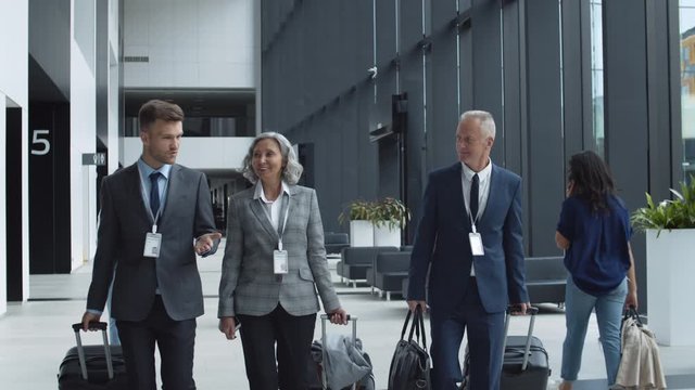 Medium shot of two Caucasian businessmen of different age and aged businesswoman with suitcases in their hands going together in airport and having discussion