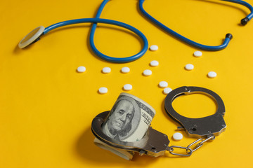 The concept of corruption in medicine. Stethoscope, pills and handcuffs with hundred dollar bills on yellow background. Medical still life. Punishment for crime