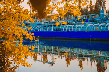 Beautiful autumn evening landscape. Colorful forest and ship quietly floating on the river. Blue barge on a background of Sunny leaves. Indian summer. Yellow trees in the rays of the departing sun