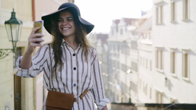 View pretty woman in hat walking at downtown of Prague. Half-length portrait girl taking selfie on smartphone, she smile and happy at background residential buildings in old district