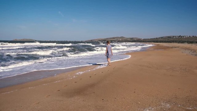 A woman walks barefoot on a sandy beach against the background of running waves and the sea on a summer day.