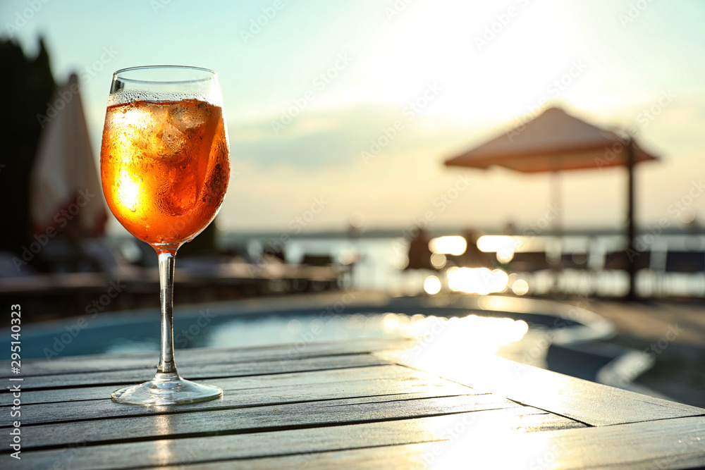 Poster Glass of fresh summer cocktail on wooden table near swimming pool outdoors at sunset. Space for text