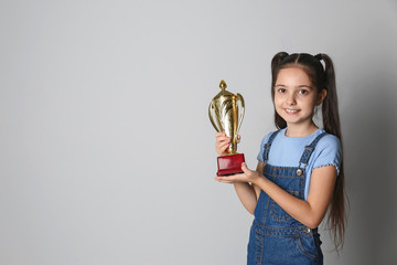 Happy girl with golden winning cup on light background. Space for text