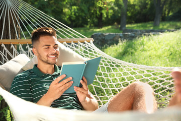 Young man reading book in comfortable hammock at green garden