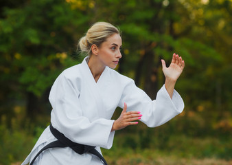 Attractive female master fighter in a white kimono with a black belt stands in a fighting stance outdoors