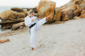 Young attractive karate woman in white kimono with a black belt trains kicks on a wild beach with stones © splitov27