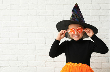 Cute little girl with candies wearing Halloween costume near white brick wall. Space for text
