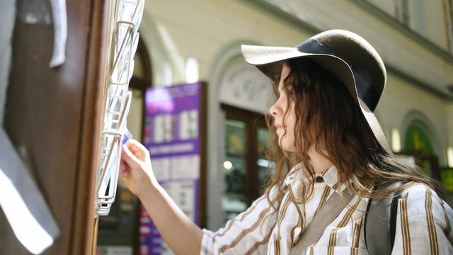 Young woman looking post cards in souvenir shop at old city centre of Prague. From below view elegant lady in hat choosing greeting cards, close-up portrait girl standing on street in backlit