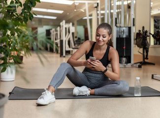 Young sport woman in sportswear using smartphone while sitting on mat in the gym. Workout break. Healthy lifestyle concept