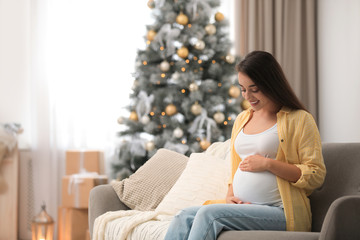 Happy pregnant woman on sofa in living room decorated for Christmas. Expecting baby