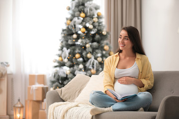 Happy pregnant woman with book in living room decorated for Christmas. Expecting baby