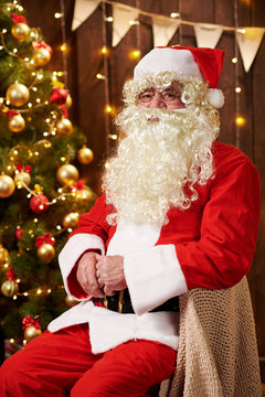 Santa Claus portrait, sitting indoor near decorated xmas tree with lights - Merry Christmas and Happy Holidays!