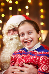 Santa Claus and child girl posing together indoor near decorated xmas tree with lights, they talking and smiling - Merry Christmas and Happy Holidays!