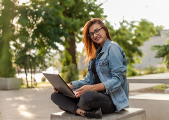 Young modern woman blogger in a denim jacket and glasses holds a laptop in his hands in the park. Online learning, blogging