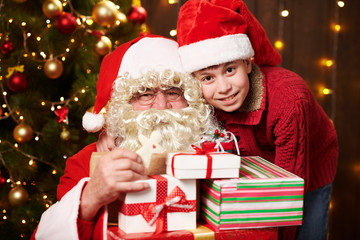 Santa Claus and santa helper boy posing with many gifts, sitting indoor near decorated xmas tree with lights - Merry Christmas and Happy Holidays!