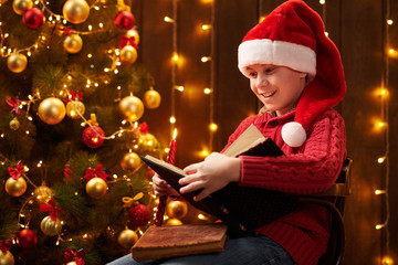 Teen boy reading book, sitting indoor near decorated xmas tree with lights, dressed as Santa helper - Merry Christmas and Happy Holidays!