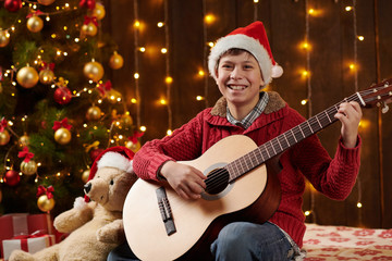 Teen boy playing guitar, sitting indoor near decorated xmas tree with lights, dressed as Santa helper - Merry Christmas and Happy Holidays!
