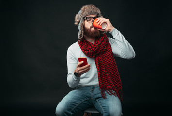 Fototapeta na wymiar Hipster with beard is drinking from a to go cup while holding and surfing on his phone.