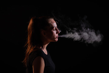 The brunette smokes an electronic cigarette on a black background, releases thick smoke from her...
