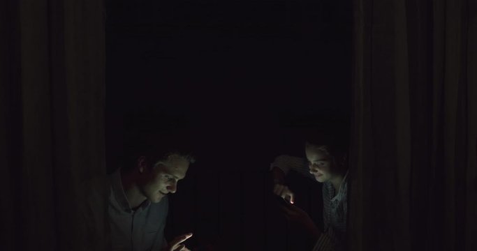 Young  Blonde Couple Swiping Smart Phone Internet Device behind Curtains at Night. Smiling Student Siblings in Evening Light Communicating with Technology, Bohemian Millennial at Twilight.