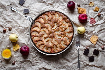 top view of the autumn dinner: Apple pie, two forks and decorations in the form of candles, apples, fallen leaves, chocolate, nuts on crumpled kraft paper