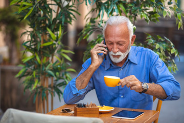Smiling happy mature man with white stylish short beard using smartphone gadget serving internet at coffee shop cafe outdoor. Laughing old man using social media network technology feeling excited.