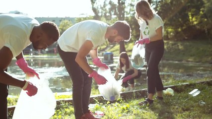 Young team of environmental volunteers picking up garbage into plastic bags and clearing polluted city park in sunny weather. Save nature. Concept of ecology. - Powered by Adobe