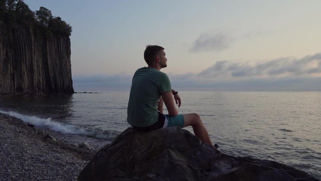 A thoughtful man sits on a rock near the sea and enjoys nature at sunset .