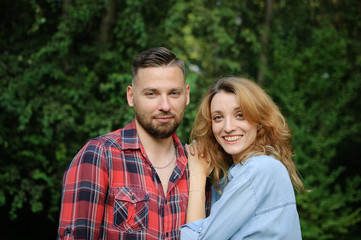 Outdoors portrait of bearded hipster boyfriend in red shirt and blond Girlfriend wearing blue casual jeans blouse in the park on green trees background