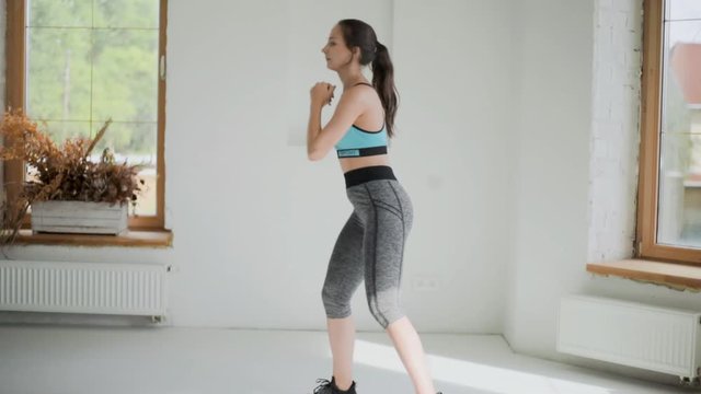 Young fit brunette girl in blue top, gray leggings, black sneakers does exercises in bright room. Doing sports at home. Healthy lifestyle. Squats, female raises and lowers her body, slow motion