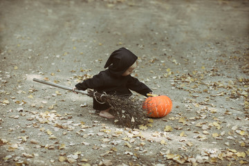 child in black halloween sorcerer or witch suit with broom sits near fresh pumpkin and carefully touches it, autumn leaves