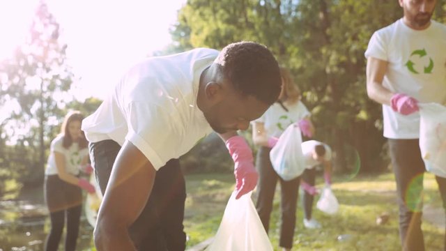 Young african activist in friendly hardworking team collecting trash waste products into bags. Group of volunteers in recycle T-shirts cleaning park environment on sunny day.